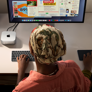 Una persona seduta a una scrivania che usa una Magic Keyboard e un Magic Trackpad per lavorare con un Mac mini collegato a uno Studio Display, accanto ci sono altri oggetti